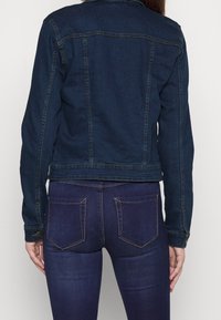 Mörkblå denimjacka med långa ärmar och klassisk krage. Har sömdetaljer och knappdekorationer. Bärs över mörkblå jeans.