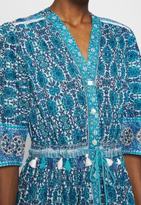 Abito floreale blu e bianco con scollatura a V, dettagli con bottoni e vita regolabile con lacci e pon pon. Maniche a sbuffo e motivi intricati.