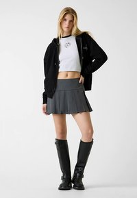 Sweat à capuche noir zippé porté sur un t-shirt blanc court, associé à une mini-jupe plissée grise et des bottes noires mi-mollet. Design simple, sans motifs.