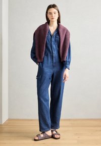 Farmernadrág, testhez sima felsőrésszel és bő szárú nadrággal, mély burgundi kötött pulóverrel a vállakon. Barna szandál.