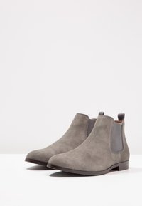 Bottes chevilles en daim gris avec panneaux latéraux élastiques, bouts arrondis et talons bas empilés. Texture lisse avec peu de détails.