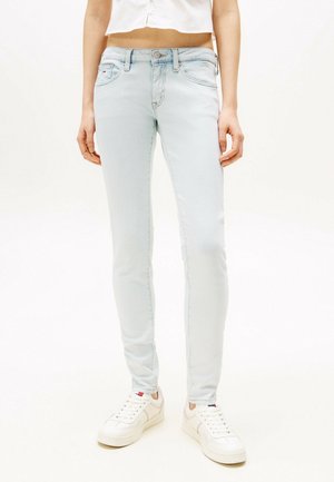 Osoba ubrana w jasnoniebieskie obcisłe dżinsy, biały krótki top i białe trampki stojąca na jednolitym tle.