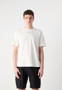 Baltas kokvilnas T-krekls ar apaļu kakla izgriezumu, īsām piedurknēm un nelielu melnu "Calvin Klein" logo krūšu centrā. Valkāts ar melniem šortiem.