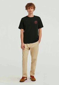Black cotton t-shirt with short sleeves, featuring a red circular logo. Paired with beige pants and brown sneakers. Model stands centered.