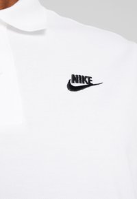 Vitt polo skjorta i texturerad tyg, med en svart broderad Nike-logotyp på övre vänstra bröstområdet.