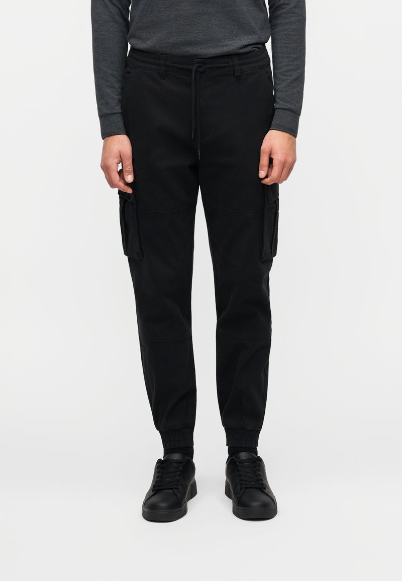 Μαύρο cargo παντελόνι σε χαλαρή εφαρμογή, με δύο πλαϊνές τσέπες, ελαστικές μανσέτες και ελαστική μέση με κορδόνι για ρύθμιση. Λείο υφή.