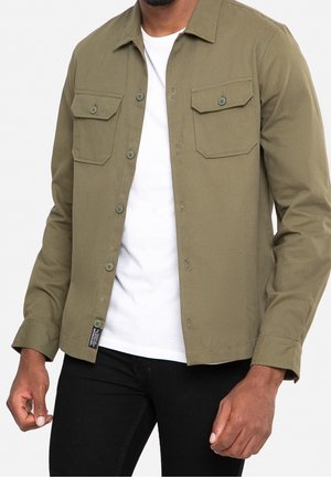Mann trägt ein offenes olivgrünes Hemd über weißem T-Shirt und schwarzen Hosen, steht mit entspannten Armen an den Seiten.