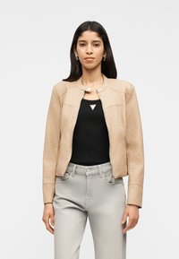 Γυναίκα με μακριά σκουρόχρωμα μαλλιά, φοράει μπεζ ζακέτα με φερμουάρ πάνω από μαύρο τοπ και ανοιχτό γκρι τζιν, στέκεται μπροστά σε απλό ανοιχτό φόντο.