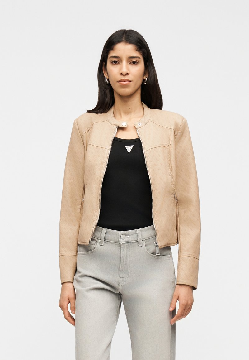 Γυναίκα με μακριά σκουρόχρωμα μαλλιά, φοράει μπεζ ζακέτα με φερμουάρ πάνω από μαύρο τοπ και ανοιχτό γκρι τζιν, στέκεται μπροστά σε απλό ανοιχτό φόντο.