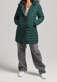 Chaqueta larga acolchada verde con capucha, adornada con un ribete de piel sintética, bolsillos con cremallera y un corte holgado, combinada con vaqueros grises de pierna ancha y zapatillas blancas.