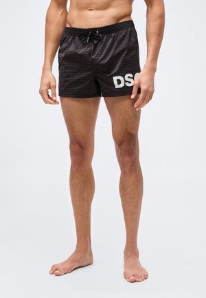Homme portant un short de bain noir court avec taille à cordon et inscription blanche "DSQ" sur la cuisse droite avant, debout pieds nus sur fond blanc.