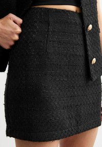 Zwarte taille Rok in tweed met een textuur, met een getailleerd silhouet en een zoom met lichte rafeling. Twee gouden knopen zichtbaar.
