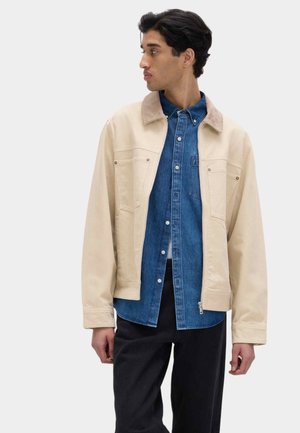 Giacca beige con design strutturato, con tasche frontali e colletto a contrasto, indossata sopra una camicia di jeans blu. Pantaloni neri.