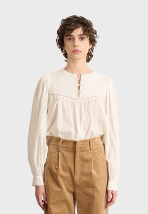 Witte textuurblouse met lange mouwen, voorzien van een ronde halslijn en een knoopsluiting met drie knopen. Gepaard met bruine corduroy broeken met hoge taille.