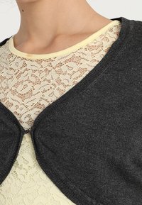 Tmavě šedý cardigan vyrobený z měkkého pleteného materiálu, s volným designem a zaoblenými okraji, vrstvený přes bledě žlutý krajkový top.
