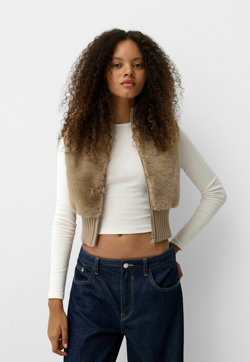 Beige Kunstfell-Cropped-Weste mit geripptem Stricksaum, getragen über einem weißen Langarmoberteil und kombiniert mit dunkelblauen High-Waist-Jeans.