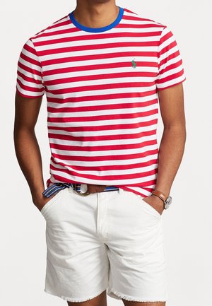 Rot-weiß gestreiftes Baumwoll-T-Shirt mit blauem Halsauschnitt, das ein grünes gesticktes Logo auf der linken Brust zeigt, kombiniert mit weißen Shorts.