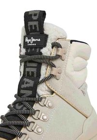 Stivale color beige in camoscio con fodera in pelliccia sintetica, lacci a trama bianca e nera e etichetta marchiata sulla linguetta. Presenti occhielli e dettagli in metallo.