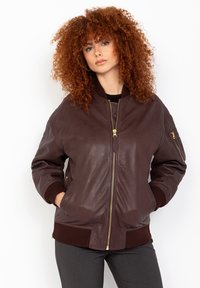 Veste bomber en cuir marron avec fermeture éclair à l'avant, poignets et ourlet côtelés, comportant des poches latérales et une poche zippée sur la manche. Texture lisse.