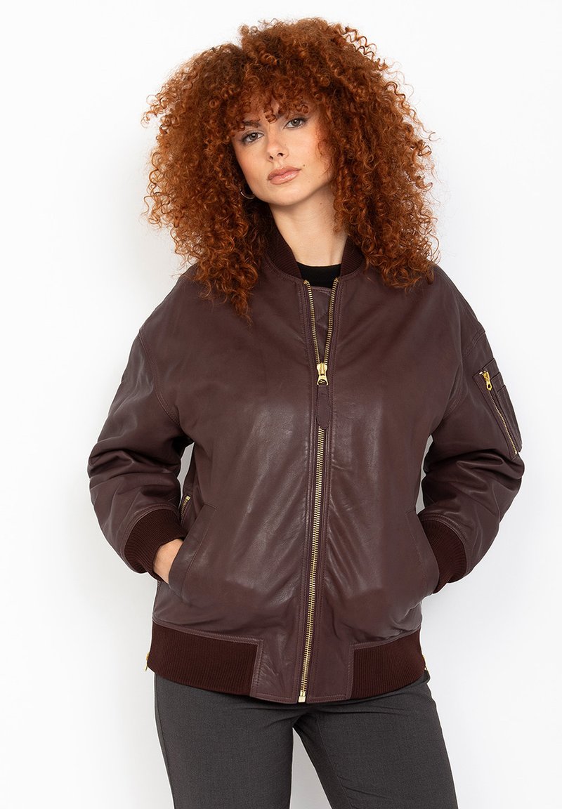 Veste bomber en cuir marron avec fermeture éclair à l'avant, poignets et ourlet côtelés, comportant des poches latérales et une poche zippée sur la manche. Texture lisse.