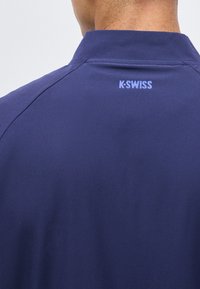 Πίσω όψη ατόμου που φοράει μπλε σκούρο πουκάμισο με το λογότυπο K-SWISS τυπωμένο κάτω από τον γιακά στο πάνω μέρος της πλάτης.