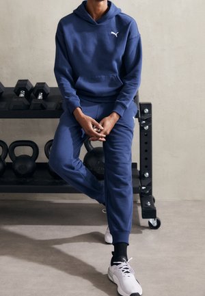 Personne en survêtement Puma bleu assise sur un banc de gym avec des haltères et kettlebells en arrière-plan, portant des baskets blanches.