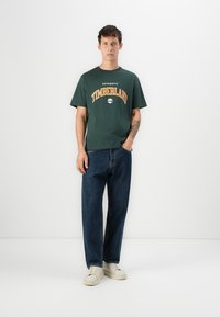 Joven de pie con una mano en el bolsillo, vistiendo una camiseta de Timberland verde oscuro, jeans azules, zapatillas crema y mostrando un tatuaje en el antebrazo.