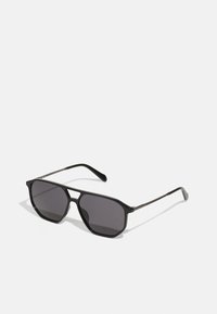 Lunettes de soleil noires avec une monture géométrique, des verres foncés et des branches métalliques fines. Le design présente une forme angulaire et une finition mate.
