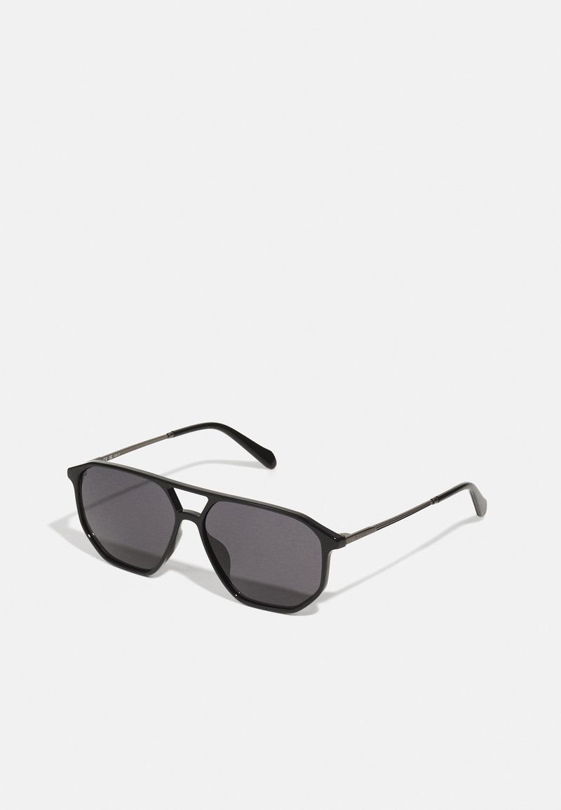 Lunettes de soleil noires avec une monture géométrique, des verres foncés et des branches métalliques fines. Le design présente une forme angulaire et une finition mate.