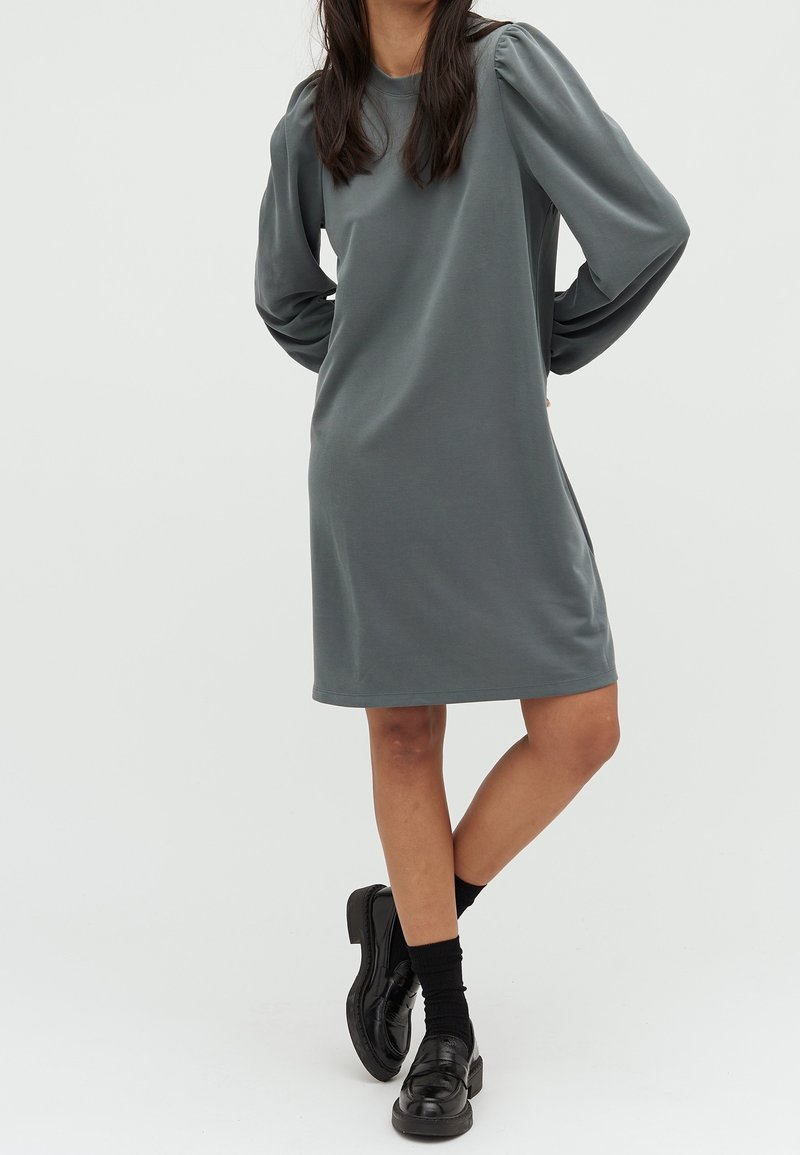 Robe grise à manches longues avec un col rond, une coupe décontractée et des épaules froncées, associée à des mocassins noirs et des chaussettes.