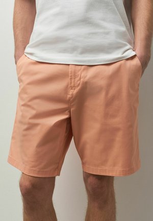 Mann, der lachsfarbene Shorts und ein weißes T-Shirt trägt, mit den Händen in den Taschen, vor einem einfarbigen hellen Hintergrund stehend.