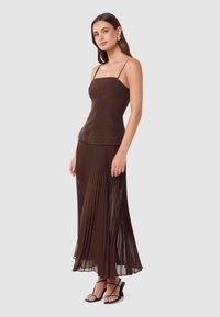 Haut en lin marron avec des bretelles spaghetti associé à une jupe plissée marron. La jupe est transparente, créant un effet superposé. Sandales noires à brides.