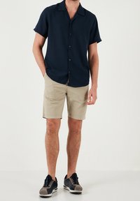 Granatowa koszula z krótkim rękawem z zapięciem na guziki, beżowe szorty oraz granatowe tenisówki z szarymi akcentami, na jednolitym tle.
