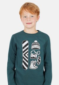 Dunkelblaues Langarmshirt mit einem weißen Grafikdesign von einem Snowboard, einer Mütze, einem Skipass und Skischuhen.