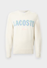 Pulover crem cu manșete și tiv cu nervuri. Prezintă logo-uri brodate multicolore „LACOSTE” și „Paris” în nuanțe pastelate delicate.
