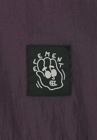 Purple fabric with a textured finish featuring a black square patch. The patch displays a white cartoon hand design with stylized letters.