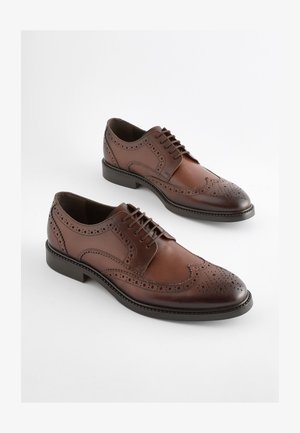 Pruunid nahast paeltega kingad, millel on brogue-detailid, sile viimistlus, ümar varvas ja astmeline konts. Tekstuursed aktsendid kõikjal.