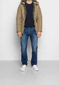 Beige puffjacka med en grön fodrad huva, marinblå stickad tröja, blå jeans och vita sneakers. Har dragkedja och fickor.