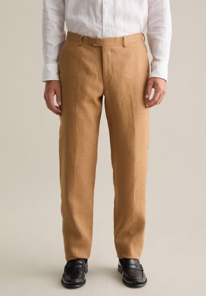 Pantalones de lino de color marrón claro con diseño de pierna recta y cierre de botón, combinados con una camisa blanca de manga larga y zapatos negros.
