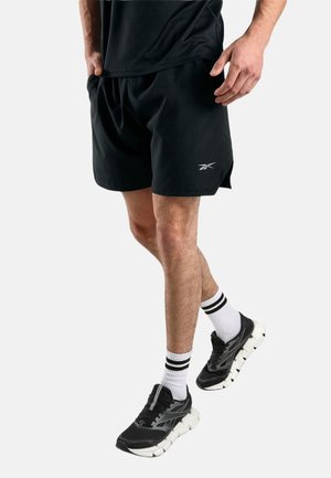 Man draagt zwarte sportshorts, zwart-witte hardloopschoenen en witte sokken met zwarte strepen, staande tegen een effen lichte achtergrond.