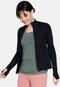 Schwarze Zip-Jacke aus glattem Stoff, mit langen Ärmeln und Daumenlöchern, über einem grün-weiß gestreiften Tanktop getragen.