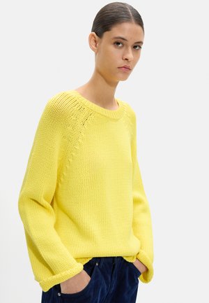 Młoda osoba związała włosy, ubrana w jaskrawożółty dziergany sweter i ciemnoniebieskie spodnie, ręce w kieszeniach, neutralne tło.