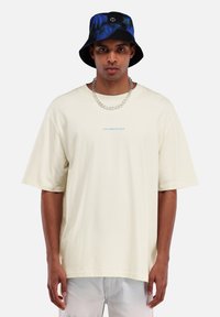 Creme, oversized t-shirt met korte mouwen en een blauwe "TOMORROWLAND" logo. Gecombineerd met een zwarte en blauwe geprinte bucket hat.