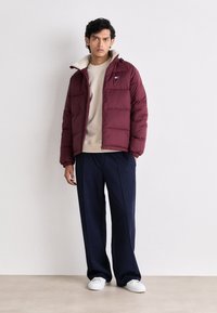 Bordeauxfarbene Daunenjacke mit einem weichen, hellen Innenfutter, ausgestattet mit einem Reißverschluss und einer aufgesetzten Tasche. Kombiniert mit navyfarbenen Hosen und weißen Sneakers.