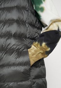 Vadderad svart väst med quiltad textur. En tie-dye ärm i grönt, svart och brunt syns från sidan, med ribbade manschetter.