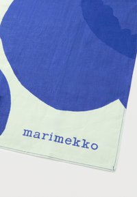 El mantel de Marimekko presenta grandes círculos azules sobre un fondo verde claro, con un logo visible en tela azul. Material de algodón texturizado.