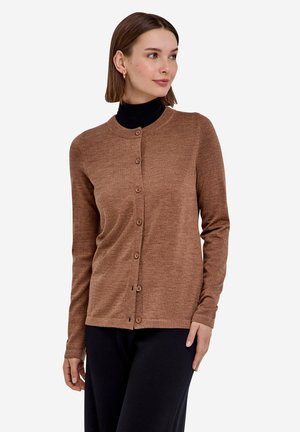 Femme aux cheveux bruns courts portant un cardigan marron boutonné par-dessus un col roulé noir et un pantalon noir, regardant légèrement sur le côté.