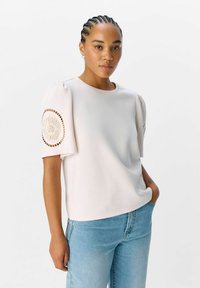 Lichtbeige top met korte mouwen en opgeblazen mouwen, versierd met geborduurde accenten op de schouders. Gepaard met lichtblauwe denim jeans.