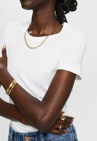 T-shirt bianco a maniche corte, scollatura rotonda, abbinato a una collana d'oro e a diversi braccialetti d'oro. Pantaloni corti in denim blu visibili in vita.
