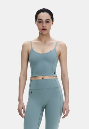 Giovane donna con capelli scuri raccolti indossa un crop top aderente azzurro chiaro e leggings a vita alta, in piedi davanti a uno sfondo chiaro e semplice.